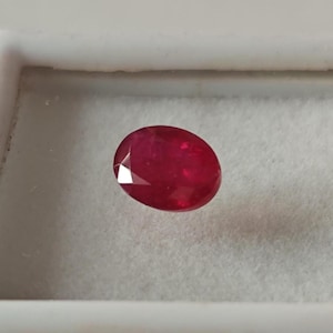 Può includere: Una gemma di rubino sfaccettata, taglio ovale. La gemma è di un colore rosso intenso e vibrante, posta su uno sfondo bianco. Le sfaccettature catturano la luce, creando un effetto scintillante. Adatta per gioielli.