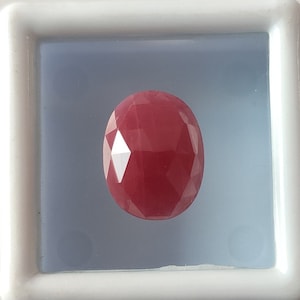 Puede incluir: Una piedra preciosa de color rojo intenso con forma ovalada facetada. La piedra preciosa se muestra en una caja blanca con un interior azul claro.