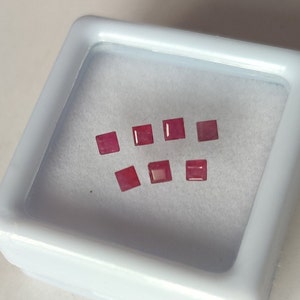 May include: A small white box with eight pink square gemstones inside.