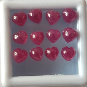 May include: Twelve heart-shaped ruby gemstones in a white display box. The gemstones are a deep red color and have a glossy finish.