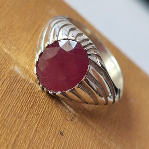 Peut inclure: Une bague en argent avec une grande pierre précieuse de rubis rouge ronde sertie au centre. La bande de la bague a un design texturé et rainuré.