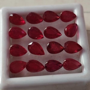 Op de afbeelding: Een verzameling van veertien peervormige, gefacetteerde robijnen. De edelstenen hebben een diepe, rijke rode kleur en zijn gerangschikt in een witte tray. De facetten vangen het licht op. Deze robijnen zijn geschikt voor het maken van sieraden.