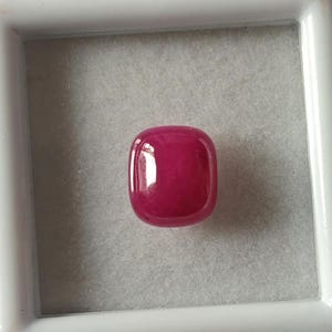 May include: A vibrant, deep pink gemstone with a smooth, rounded, cushion-shaped cut. The gemstone is displayed in a white square box.