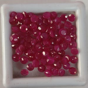 May include: A collection of small, round, faceted, vibrant pinkish-red gemstones arranged closely together within a square white display case. Each individual stone features multiple reflective surfaces that catch the light, creating a uniform appearance across the group of loose, polished, circular gems.