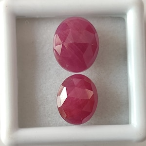 Op de afbeelding: Twee ovale, gefacetteerde robijn edelstenen in een witte doos. De robijnen zijn diep roze.
