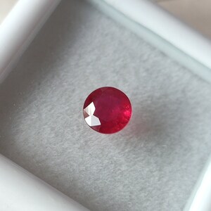 Peut inclure: Une pierre précieuse ronde et rouge avec des facettes. La pierre précieuse est placée sur un fond blanc.