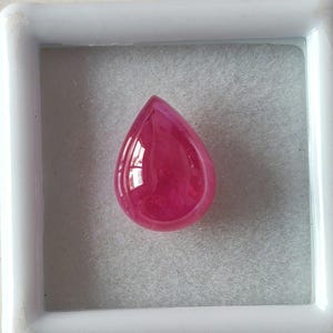 Può includere: Una pietra preziosa rosa a forma di lacrima con una superficie liscia e lucida. La pietra preziosa è esposta in una scatola bianca.