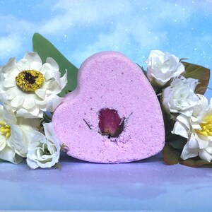 May include: A heart-shaped, light pink bath bomb with a dried rose bud in the center. The bath bomb is surrounded by white flowers with yellow centers and green leaves, set against a blue background.