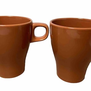 May include: Two orange-brown ceramic mugs with curved sides and attached handles. The mugs have a glossy finish and a simple, modern design, suitable for coffee or tea.