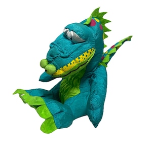 May include: A teal plush dragon toy with green and yellow accents. The dragon has a whimsical design with a large head, expressive eyes, and a playful pose. The dragon's back features green spikes and pink and yellow spots.