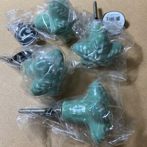 May include: A collection of green ceramic cabinet knobs in the shape of birds, packaged in clear plastic. One knob has a black and white design. The knobs have metal screws for installation.