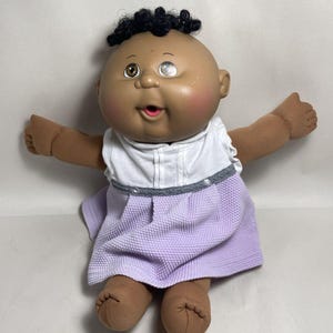 May include: A doll with dark skin, black curly hair, and heterochromia, featuring one brown eye and one light blue eye. The doll wears a white sleeveless top with a textured lavender skirt, pink cheeks, and an open mouth expression with a pink interior.