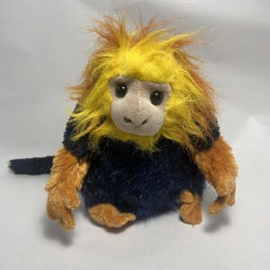 May include: A plush toy monkey with a yellow and orange furry head, a tan face, and dark eyes. The monkey has a navy blue body and tan arms and feet. The tail is navy blue. The toy is designed to resemble a golden lion tamarin.