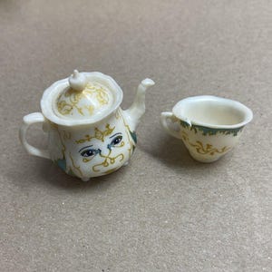 May include: A miniature cream-colored teapot and teacup set. The teapot has a lid and is decorated with gold and blue accents, including a face design. The teacup has a handle and matching gold and green floral details.