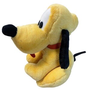 Pode incluir: Um peluche de cão com pelo amarelo, um nariz preto grande, orelhas pretas compridas e uma coleira vermelha. A personagem tem manchas brancas à volta dos olhos e uma pequena língua vermelha a sair da boca, em posição sentada.