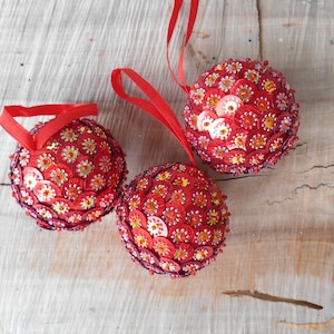 May include: Three red Christmas ornaments with sequin and bead embellishments. Each ornament is spherical, with a red ribbon loop for hanging. The ornaments are decorated with red sequins and small yellow and orange beads.