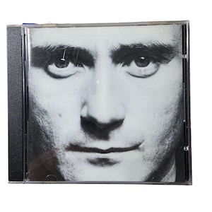 May include: A square compact disc jewel case featuring a black and white close-up portrait of a person's face on the front cover. The case has a black plastic spine and a clear plastic front panel, displaying the high-contrast facial features.