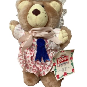 May include: A plush brown teddy bear wearing a floral dress with a blue ribbon and a lace bonnet. The bear has a cream-colored snout and a tag that reads "Wendy's Hamburgers".