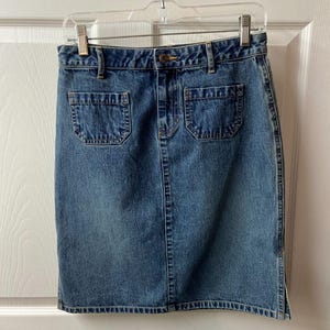 May include: A denim pencil skirt in a medium wash blue. The skirt has two front pockets, a button closure, and a small slit at the back. The skirt is hanging on a clear plastic hanger.