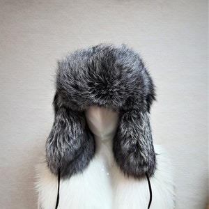 May include: A silver fox fur hat with ear flaps and a matching white fox fur collar. The hat and collar are tied together with a black cord.