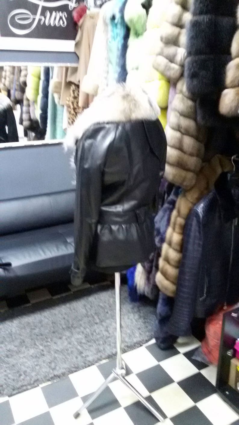 Pu&ograve; includere: Una giacca di pelle nera con colletto di pelliccia e cintura. La giacca &egrave; su un espositore in un negozio.
