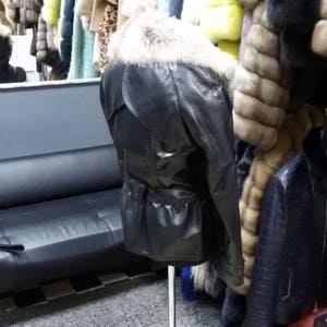 Pu&ograve; includere: Una giacca di pelle nera con colletto di pelliccia su un manichino in un negozio di pellicce. La giacca ha una cintura e una chiusura a zip. Il negozio &egrave; decorato con un pavimento a scacchi e un tappeto grigio.