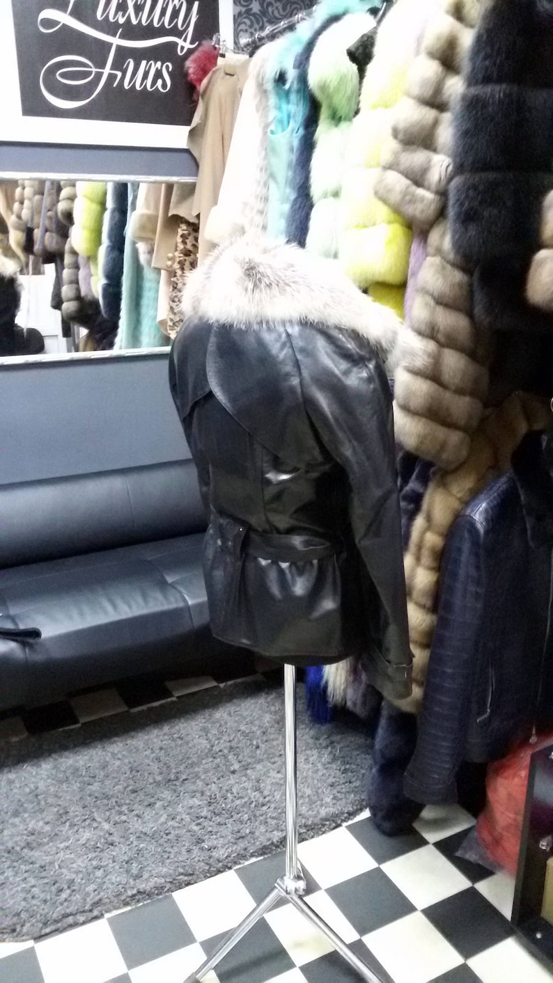 Pu&ograve; includere: Una giacca di pelle nera con colletto di pelliccia su un manichino in un negozio di pellicce. La giacca ha una cintura e una chiusura a zip. Il negozio &egrave; decorato con un pavimento a scacchi e un tappeto grigio.