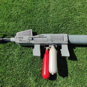 Könnte beinhalten: Eine graue Requisitenpistole mit zylindrischem Lauf und Griff. Unten sind eine rote und eine weiße Kartusche befestigt. Die Requisite befindet sich vor einem grünen, grasbewachsenen Hintergrund.