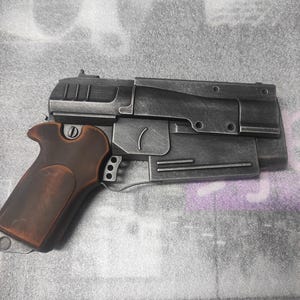 Puede incluir: Una pistola de utilería gris metálico y marrón con un diseño detallado. El arma presenta una empuñadura marrón y un cuerpo gris con varios componentes mecánicos. La estética general sugiere un tema futurista o de ciencia ficción.