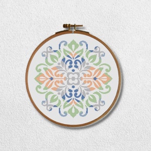 Peut inclure: Un cerceau de broderie au point de croix avec un fond blanc et un motif géométrique coloré dans des tons de bleu, vert et pêche. Le motif est un dessin symétrique avec un motif floral.