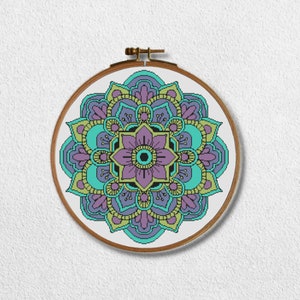 Könnte beinhalten: Ein farbenfrohes Mandala-Muster in Violett-, Türkis- und Gelbtönen ist auf eine weiße Leinwand gestickt. Die Leinwand ist auf einem hölzernen Stickrahmen gespannt.