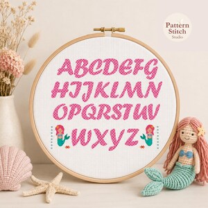 Puede incluir: Un bastidor de bordado circular de madera que contiene tela Aida blanca con un alfabeto de punto de cruz en letras rosas estampadas. El diseño incluye dos pequeñas figuras de sirenas y dos cangrejos flanqueando las letras W a Z abajo.