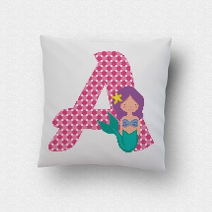 Puede incluir: Un cojín cuadrado blanco presenta la letra "A" rosa con un estampado de cuadros. Una sirena de dibujos animados con cabello morado, un top azul y una cola verde está en el lado derecho de la letra. Una estrella amarilla está en su cabello.
