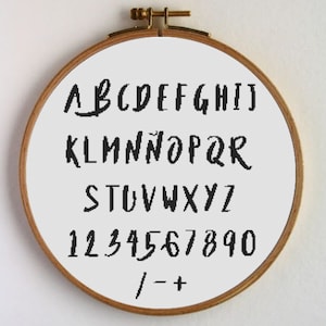 Puede incluir: Un aro de bordado blanco con letras y números escritos a mano en negro. Las letras son de la A a la Z y los números del 1 al 9 y el 0. Los símbolos / - + también están incluidos.