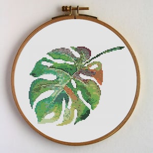 Puede incluir: Un aro de bordado de punto de cruz con un diseño de hoja de monstera verde, amarilla y naranja.