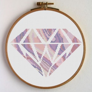 Puede incluir: Un patrón de punto de cruz de un diamante rosa y morado con un contorno blanco. El diamante está en un aro de bordado de madera.