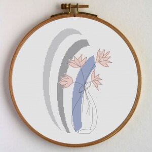 Puede incluir: Bordado de punto de cruz con un diseño floral minimalista. La obra de arte presenta un jarrón con flores rosas y una franja vertical azul sobre un fondo blanco. El bordado está enmarcado en un aro de madera redondo.