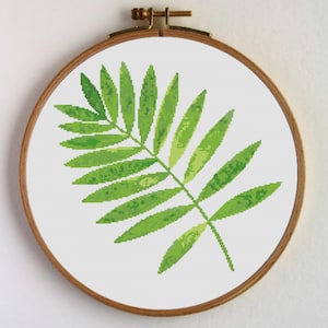 Puede incluir: Una rama verde con hojas con efecto acuarela, bordada en punto de cruz y expuesta en un aro de bordado de madera.