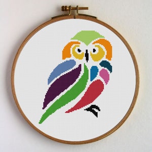 Puede incluir: Un patrón de punto de cruz geométrico colorido de un búho en un aro de bordado de madera. El búho está formado por diferentes formas de colores, incluyendo verde, rojo, morado, azul y amarillo.