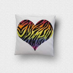 Puede incluir: Una almohada blanca con un diseño en forma de corazón de colores. El corazón es un estampado de cebra arcoíris con rayas negras y un esquema de colores rojo, naranja, amarillo, verde, azul y morado.