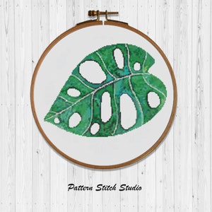 Puede incluir: Un patrón de punto de cruz de una hoja de monstera verde en un aro de bordado de madera. La hoja tiene un efecto acuarela con tonos de verde y azul.