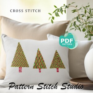 Puede incluir: Una almohada blanca con tres árboles de Navidad coloridos bordados en punto de cruz. La almohada está sobre un sofá beige con una planta en el fondo. El texto "CROSS STITCH" está en la parte superior de la imagen y "Pattern Stitch Studio" en la parte inferior. Un círculo verde y blanco con el texto "PDF PATTERN" está en la esquina superior derecha.