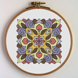 Puede incluir: Un patrón de punto de cruz colorido con un diseño de mandala amarillo, marrón, morado y rosa. El patrón está enmarcado en un aro de bordado de madera.
