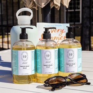 May include: Three clear glass bottles of hand and body wash with a white label and black pump tops. The label has the text "H abwond Hand & Body Wash" on it. The bottles are sitting on a white table with a pair of brown sunglasses.
