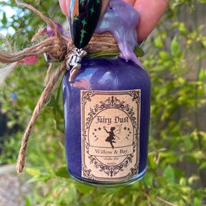 May include: A small glass bottle filled with purple powder, labelled "Fairy Dust" with a fairy illustration. The bottle is sealed with purple wax and decorated with a small fairy charm and twine. The text "Willow & Bay" and "shake the magic" are also visible.