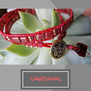 May include: A red and gold beaded wrap bracelet with a decorative gold button. The bracelet is on a green succulent plant. The text "CARDINAL" is written in red on a white rectangle.