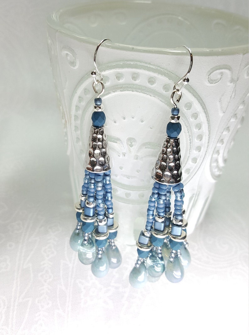 May include: A pair of silver and blue beaded tassel earrings. The earrings feature a silver cone-shaped bead at the top, followed by a cascade of blue beads and a cluster of iridescent blue teardrop-shaped beads.