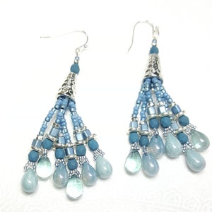 May include: A pair of dangle earrings with a silver cone-shaped top and a tassel of blue beads. The tassel is made of different shades of blue beads, including light blue, medium blue, and dark blue. The earrings are finished with a silver hook.