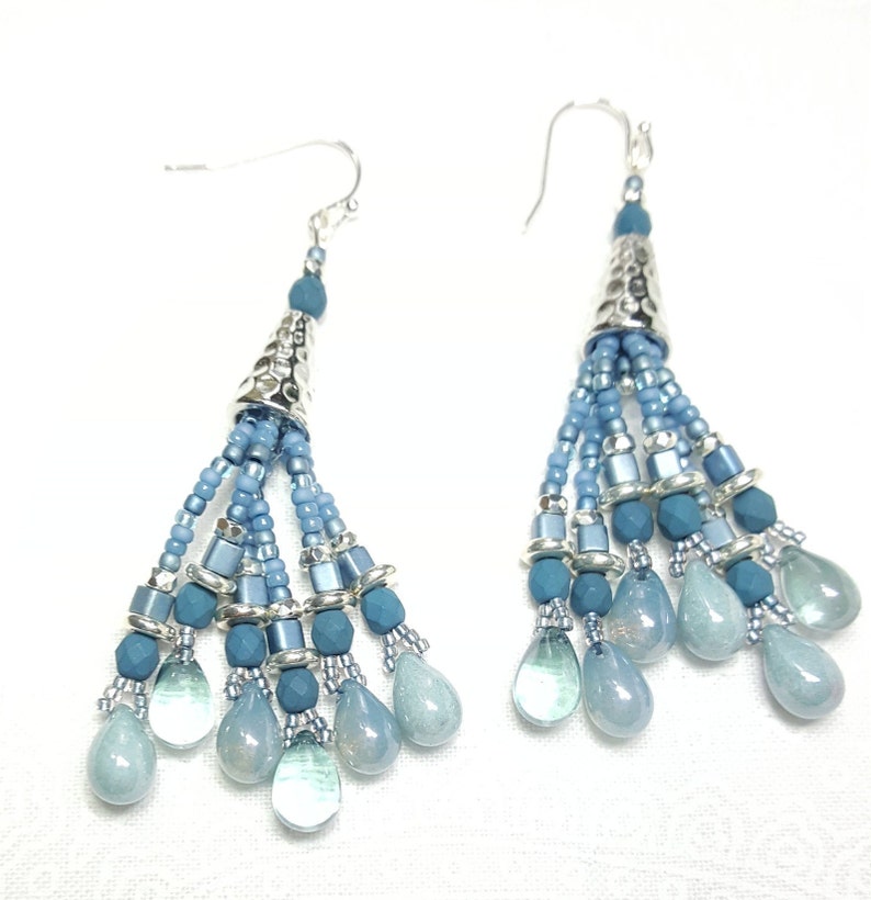 May include: A pair of dangle earrings with a silver cone-shaped top and a tassel of blue beads. The tassel is made of different shades of blue beads, including light blue, medium blue, and dark blue. The earrings are finished with a silver hook.