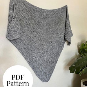 May include: A grey knitted shawl or scarf hanging on a white wall. The text "PDF Pattern" is written in a white circle on the wall.